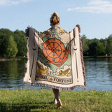 Person holding a 'Wheel of Fortune' tarot card-themed blanket by a lake.