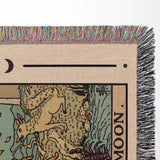 Close-up of a woven blanket with fringe, featuring a stylized illustration of a tarot card.