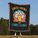 Person holding a black blanket with ghost design and text 'Ghost Shows and Comfy Clothes' against a blue sky and trees background.