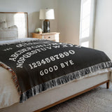 Black Ouija blanket with white text on a bed in a bedroom setting
